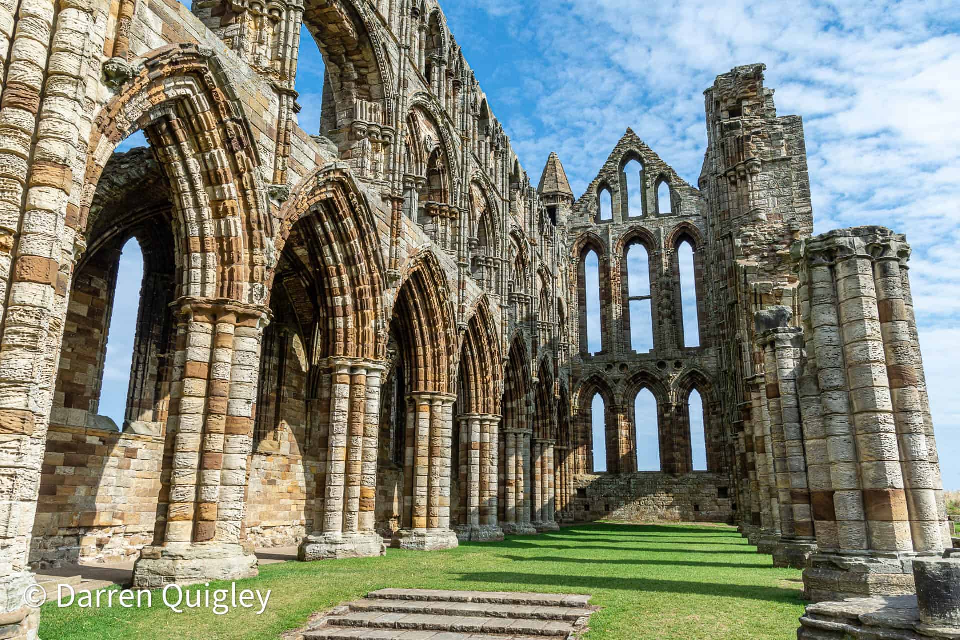 Whitby Abbey Ancient stone ruins of a historic Gothic church or abbey with tall arches and intricate stonework, under a bright blue sky with clouds, located in the UK.
